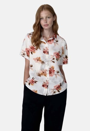 Giovane donna con lunghi capelli rossi che indossa una camicia bianca a maniche corte con motivi floreali rossi e marroni e pantaloni neri.