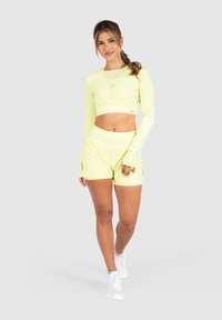 Neon-gelbes sportliches Crop-Top mit langen Ärmeln und passender Shorts. Leichter, elastischer Stoff mit Logodetail und lockerer Passform. Weiße Sneakers.
