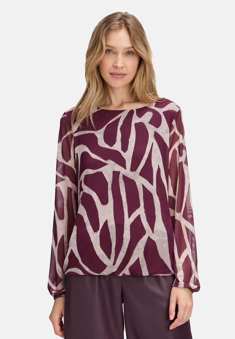 Kobieta w przezroczystej bluzce z długim rękawem w abstrakcyjny fioletowo-beżowy wzór i dopasowanych fioletowych spodniach, zwrócona przodem.