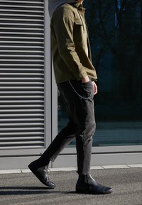 Hombre caminando afuera, vistiendo botas negras, jeans negros con una cadena y una chaqueta verde oliva con una capa con capucha gris debajo.