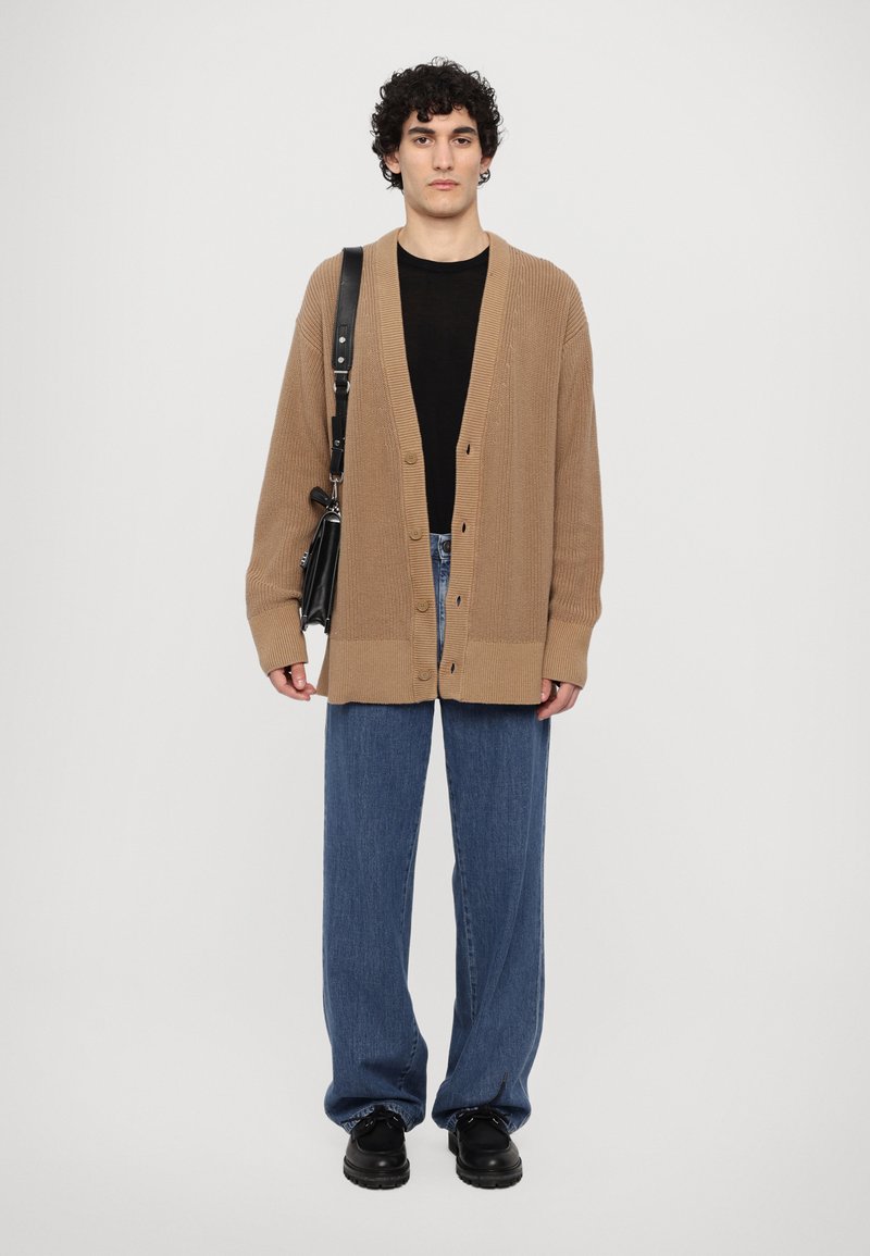 Brauner Strickcardigan mit V-Ausschnitt und Knopfverschluss, kombiniert mit einem schwarzen Top und lockeren blauen Jeans, abgerundet mit schwarzen Schuhen.