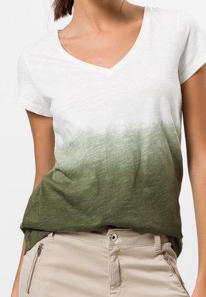 Vrouw draagt een T-shirt met korte mouwen en V-hals met een witte naar groene gradient en beige broek met ritszakken.