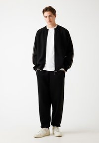 Schwarze Zip-Up-Sweatjacke mit weißen Streifen, kombiniert mit schwarzen Jogginghosen und weißen Turnschuhen. Das Model posiert vor einem schlichten Hintergrund.