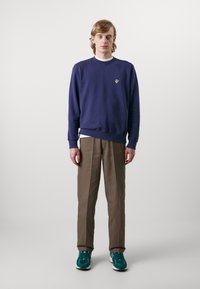 Felpa navy con polsini a coste, pantaloni beige e sneaker teal. La felpa presenta un piccolo logo sul petto. Design semplice e informale.