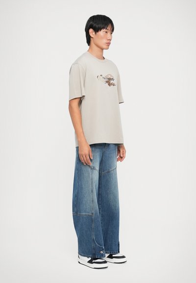 Lys grå bomull t-skjorte med grafisk trykk foran, kombinert med vide blå denimjeans med synlige sømmer og store lommer.