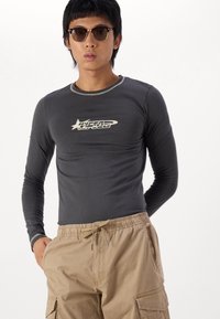 Long-sleeve gray fitted shirt with contrasting black collar, featuring a white logo graphic on the chest. Paired with beige cargo shorts.