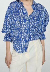 Blue floral shirt with white botanical patterns, featuring a button-up design and ruffled sleeves. Worn over light-colored trousers.