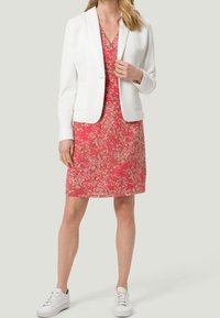 Valkoinen bleiseri vaaleanpunaisen kukkamekon päällä, jossa on lyhyet hihat, V-pääntie ja polvien ylle ulottuva helma. Valkoiset lenkkarit täydentävät asua.