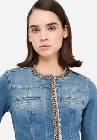 Jeans jakna svetlo modre barve z raztrganim izrezom in sprednjimi žepi, okrašena z zlato verigo po sredini.