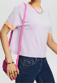 Krótki sweter w jasnofioletowym kolorze z kontrastującymi pomarańczowymi mankietami, zestawiony z ciemnoniebieskimi dżinsami i małą różow�ą torebką na ramię.