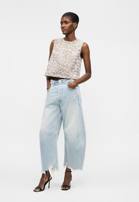 Γυναίκα στέκεται φορώντας αμάνικη μπλούζα με λεοπάρ σχέδιο, ανοιχτό μπλε φαρδιά σκισμένα τζιν και μαύρα σανδάλια με τακούνι και λουράκια στον αστράγαλο.