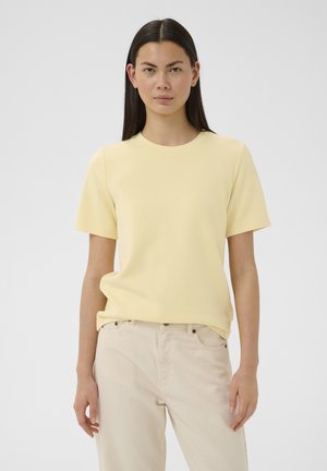 Žena s dlhými tmavými vlasmi v jednoliatom svetložltom tričku s krátkym rukávom a béžových nohaviciach, stojaca pred jednoduchým bielym pozadím.