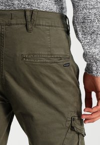 Olijfgroene cargobroek met een textuurafwerking, voorzien van een zijvak en een zwart merkje op de achterkant. De tailleband heeft riemlussen.
