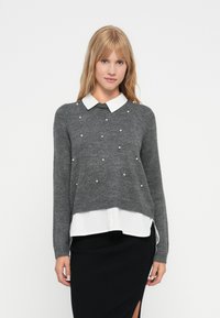 Pulover gri din tricot cu guler alb, cu accente de perle. Stratificat peste o bluză albă, asortat cu o fustă neagră cu dungi.