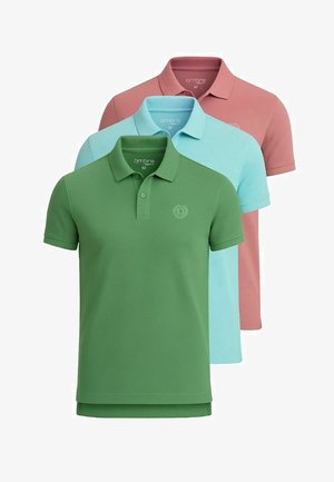 Chemise polo rose à manches courtes avec logo, associée à un pantalon cargo beige clair et des baskets blanches avec des accents marron, se tenant devant un fond blanc.