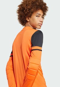 Oranje doelmanstrui met zwarte mouwen; heeft gewatteerde ellenbogen en schouderdetails; gemaakt van een gladde, rekbare stof.