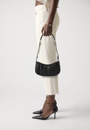 Black quilted handbag with a chain strap, held by a person wearing cream pants and black pointed-toe heels. Elegant design with hardware accent.
