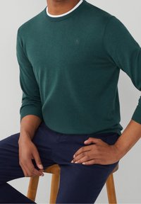 Pull vert en tricot avec encolure ronde, poignets côtelés et un logo discret sur la poitrine. Porté avec un pantalon bleu marine, assis sur un tabouret en bois.