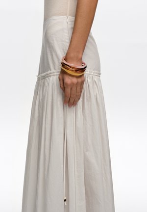 Perfil lateral de mujer con una falda blanca de cintura alta con detalles fruncidos, complementada con tres pulseras gruesas en la muñeca.