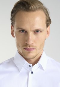 Biała koszula z wyostrzeniem, z niebieskim guzikiem przy kołnierzy. Gładka tekstura i czysty, dopasowany krój.