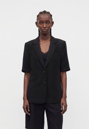 Personne grande aux cheveux tressés portant un blazer noir à manches courtes sur un haut orné de dentelle et un short noir assorti, debout devant un fond uni.