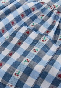 Tissu vichy bleu et blanc avec des motifs de cœurs rouges et de petits motifs floraux brodés, présentant une texture subtile et des éléments de design froncés.