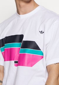 Bílý bavlněný tričko s velkým grafickým designem v růžových, černých a tyrkysových tvarech. Malé černé logo na levém rameni.