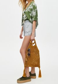 Netz-Einkaufstasche in kräftigem Gelb mit zwei Griffen und Quasten. Modell trägt ein grünes Blumenshirt und graue Shorts, dazu grüne Schuhe.