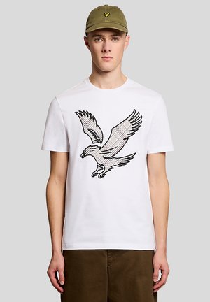 Jeune homme portant un T-shirt blanc avec un grand aigle en motif à carreaux, une casquette vert olive avec un logo jaune, et un pantalon marron.