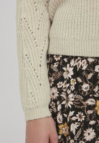 Cremefarvet strikket sweater med en struktureret vævning og ribbede manchetter, parret med en sort blomsterskirt med hvide og gule mønstre.