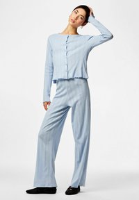 Γυναίκα που φοράει φωτεινή γαλάζια ριγωτή ζακέτα με κουμπιά και ταιριαστό παντελόνι με φαρδιά πόδια, ποζάροντας με το ένα χέρι στο κεφάλι της μπροστά σε λευκό φόντο.