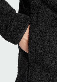 Giacca in pile nera con superficie testurizzata, che presenta una tasca laterale e bordi a costine lungo l'apertura della manica per uno stile aderente.