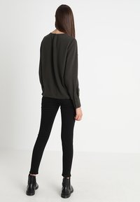Blusa de manga larga en verde oscuro, de corte holgado y con puños abotonados, combinada con pantalones ajustados negros y botas de tobillo. Textura de tela suave.