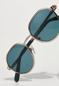 Occhiali da sole in metallo color oro con grandi lenti color teal, forma quadrata e un ponte decorativo. Presentano naselli trasparenti e astine di colore scuro.