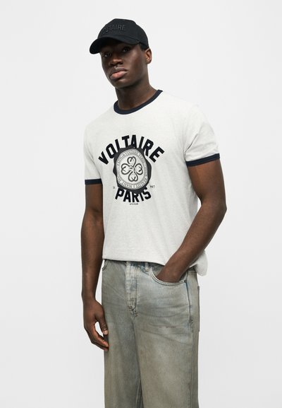 Νεαρός άνδρας που φοράει λευκό T-shirt με το κείμενο "Voltaire Paris", μαύρο καπέλο και ανοιχτό μπλε τζιν, στέκεται με το ένα χέρι στην τσέπη.