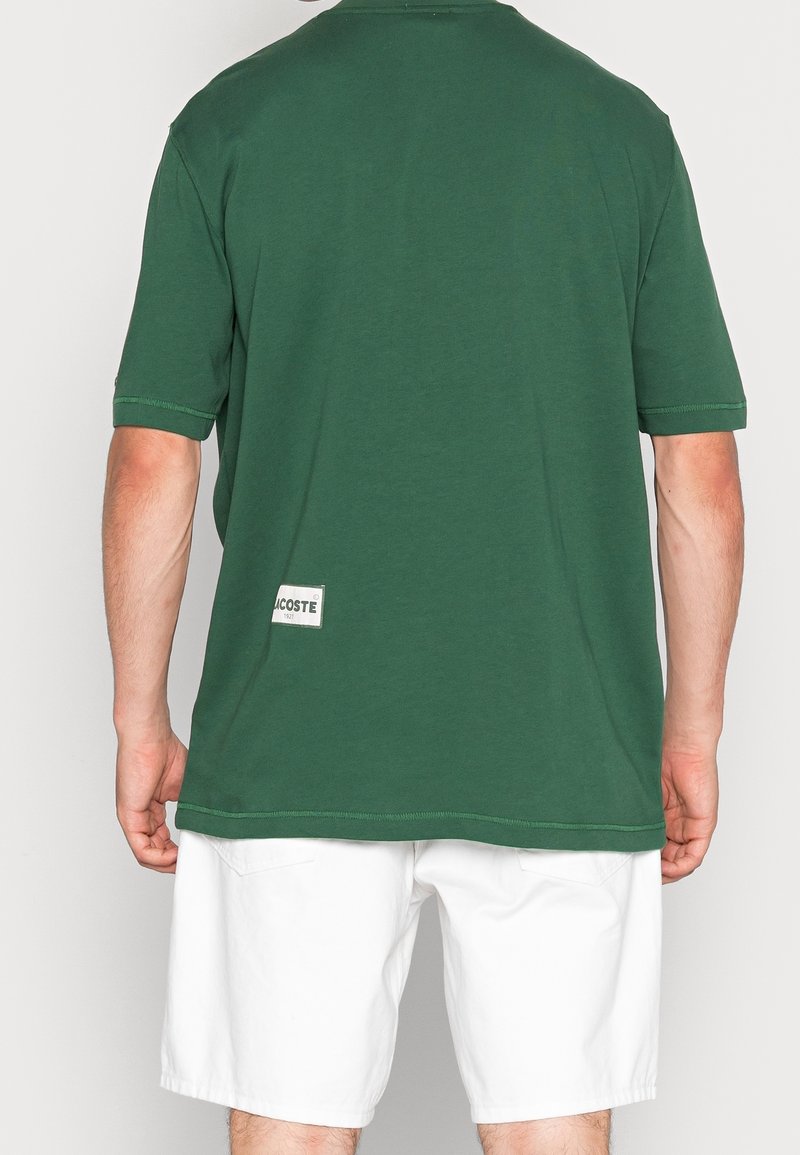 Camiseta de algodón de manga corta en color verde bosque, con corte recto y una etiqueta con "Lacoste" en la parte inferior izquierda.