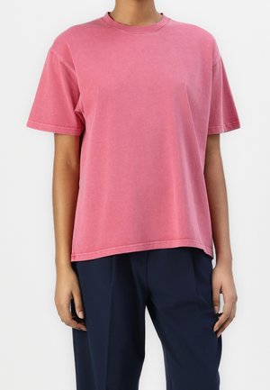 Osoba ubrana w luźny, krótki rękaw blady różowy t-shirt w wyblakłym kolorze, zestawiony z ciemnoniebieskimi spodniami, stojąca na jednolitym białym tle.