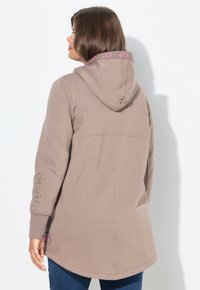 Giacca beige con cappuccio e vestibilità rilassata. Presenta accenti del logo rosa lungo il cappuccio e testo ricamato sulla manica. Tessuto morbido e testurizzato.
