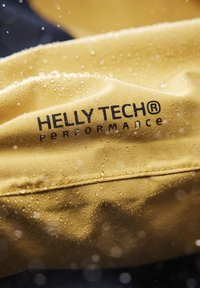 Tessuto giallo impermeabile con gocce d'acqua, che presenta il testo "HELLY TECH® PERFORMANCE" in un carattere bold e scuro.