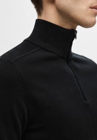 Czarny sweter z wysokim kołnierzem na zamek, z prążkowanym kołnierzem i miękką, teksturowaną tkaniną. Dopasowany, z subtelnymi szwami na ramionach.