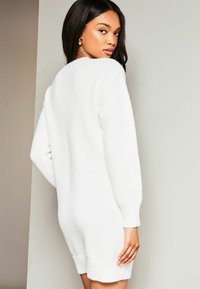 Rochie tricotată albă cu un model texturat, mâneci lungi și manșete cu nervuri. Prezintă o croială lejeră și lungime până la mijlocul coapsei.