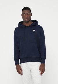 Νεαρός άνδρας που φοράει σκούρο μπλε φούτερ με τσέπη μπροστά και λευκό λογότυπο, σε συνδυασμό με ανοιχτό γκρι φόρμα, στέκεται μπροστά σε ένα απλό φόντο.