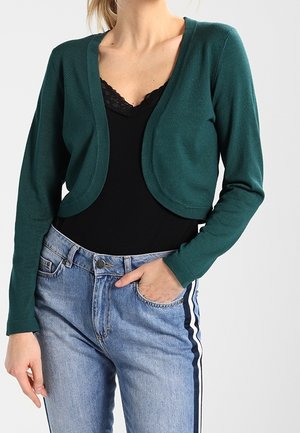 Femme portant un cardigan court vert foncé sur un top noir avec bordure en dentelle, associé à un jean bleu clair avec des bandes latérales.