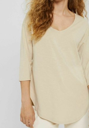 Femme aux cheveux bouclés portant une chemise beige à manches longues et col en V, ainsi qu'un pantalon blanc sur fond uni.