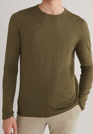 Hombre con suéter de manga larga color verde oliva y pantalones beige, con una mano en el bolsillo contra un fondo liso.