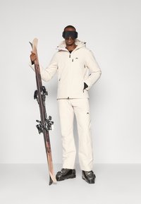 Hvit snøjakke med hette og glidelås, matchende hvite bukser, svarte skibriller, holder på ski med bindinger og svarte skistøvler.