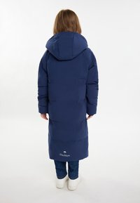 Lungo cappotto puffer blu navy con cappuccio, caratterizzato da una texture liscia, polsini elasticizzati e un dettaglio con logo vicino all'orlo. Indossato sopra a dei jeans.