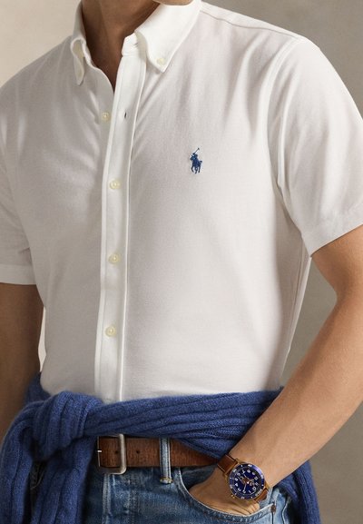 Polo branca de manga curta com um logótipo azul bordado. Combinada com malha azul amarrada à cintura e um cinto de couro castanho.