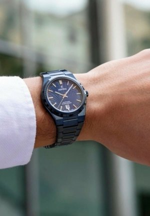 Blaues Edelstahl-Armbanduhr mit strukturiertem Zifferblatt, Datumsfenster auf 6 Uhr und Gliederarmband, auf einer Hand nahe einem weißen Hemd präsentiert.