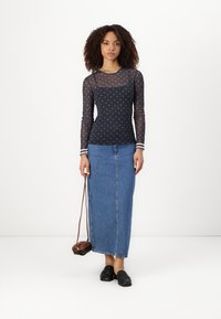 Navy long-sleeve mesh top met een subtiele print, gecombineerd met een lichtblauwe denim maxi-rok. Model houdt een kleine bruine schoudertas vast.